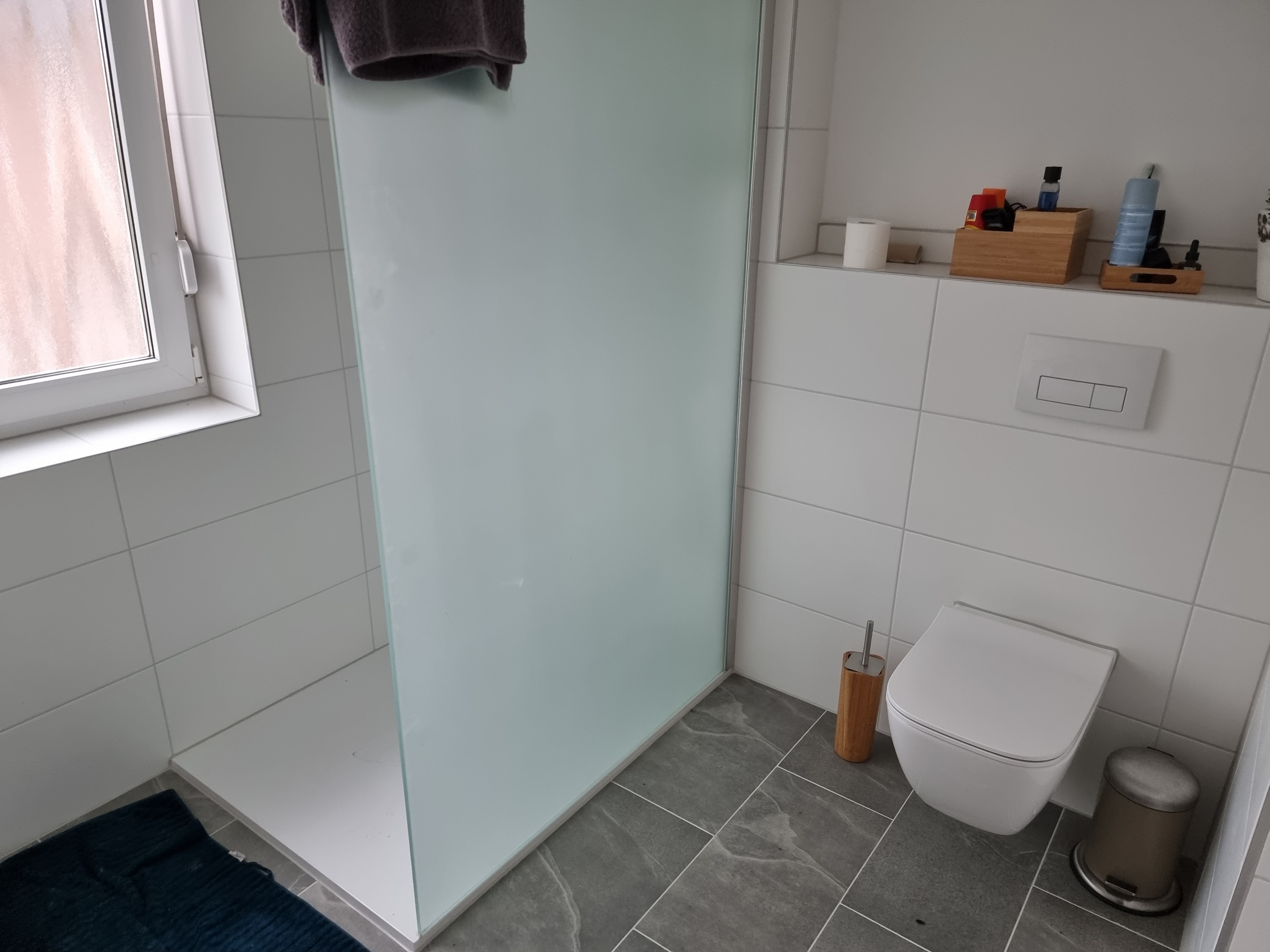 The bathroom was completely renovated by Guido Breitbach. The picture shows a bathroom with a toilet gray floor tiles and white rectangular large tiles on the wall. You can also see the shower
